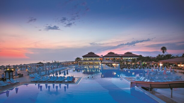 Ein Resort-Poolbereich mit Liegestühlen und Sonnenschirmen, ein großes Schwimmbecken und ein Gebäude mit mehreren Dächern.