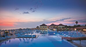 A resort pool area with lounge chairs and umbrellas, a large pool, and a building with multiple roofs. - Club Nena - All Inclusive (Manavgat)
