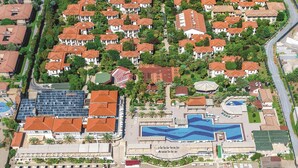 Eine Vogelperspektive zeigt einen Resortkomplex mit mehreren Gebäuden, einem großen Swimmingpool und einem Strandbereich mit Liegestühlen.