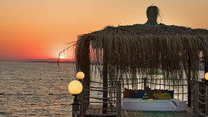 Ein Gebäude mit einem Reetdach, mit Bett und Kissen, mit Blick auf ein Gewässer bei Sonnenuntergang.
