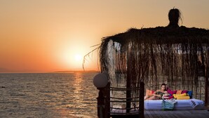 Ein Strandbett mit einem Reetdach, einer Holzterrasse und Blick auf den Ozean bei Sonnenuntergang.