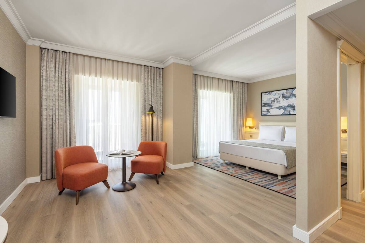 A hotel room with a bed, two armchairs, a small table, a TV, and a painting on the wall.