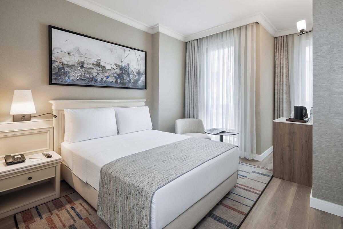 A hotel room with a large bed, a bedside table with a lamp, a chair, and a small table with a cup and a newspaper.