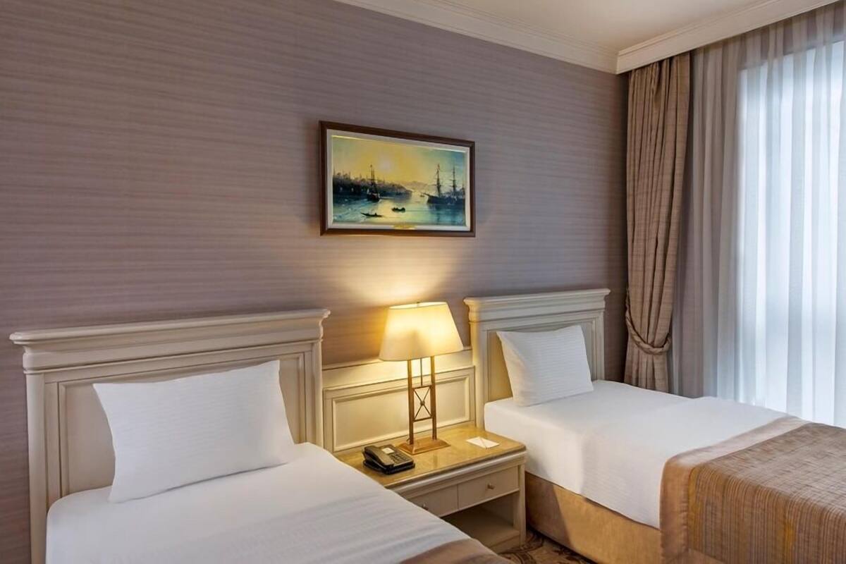 A hotel room with two beds, a nightstand with a lamp, a painting on the wall, and a window with curtains.