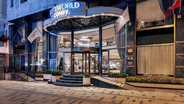 Entrada de un hotel con fachada curva, puertas de vidrio y un letrero que dice 'ELITEWORLD COMFY'.