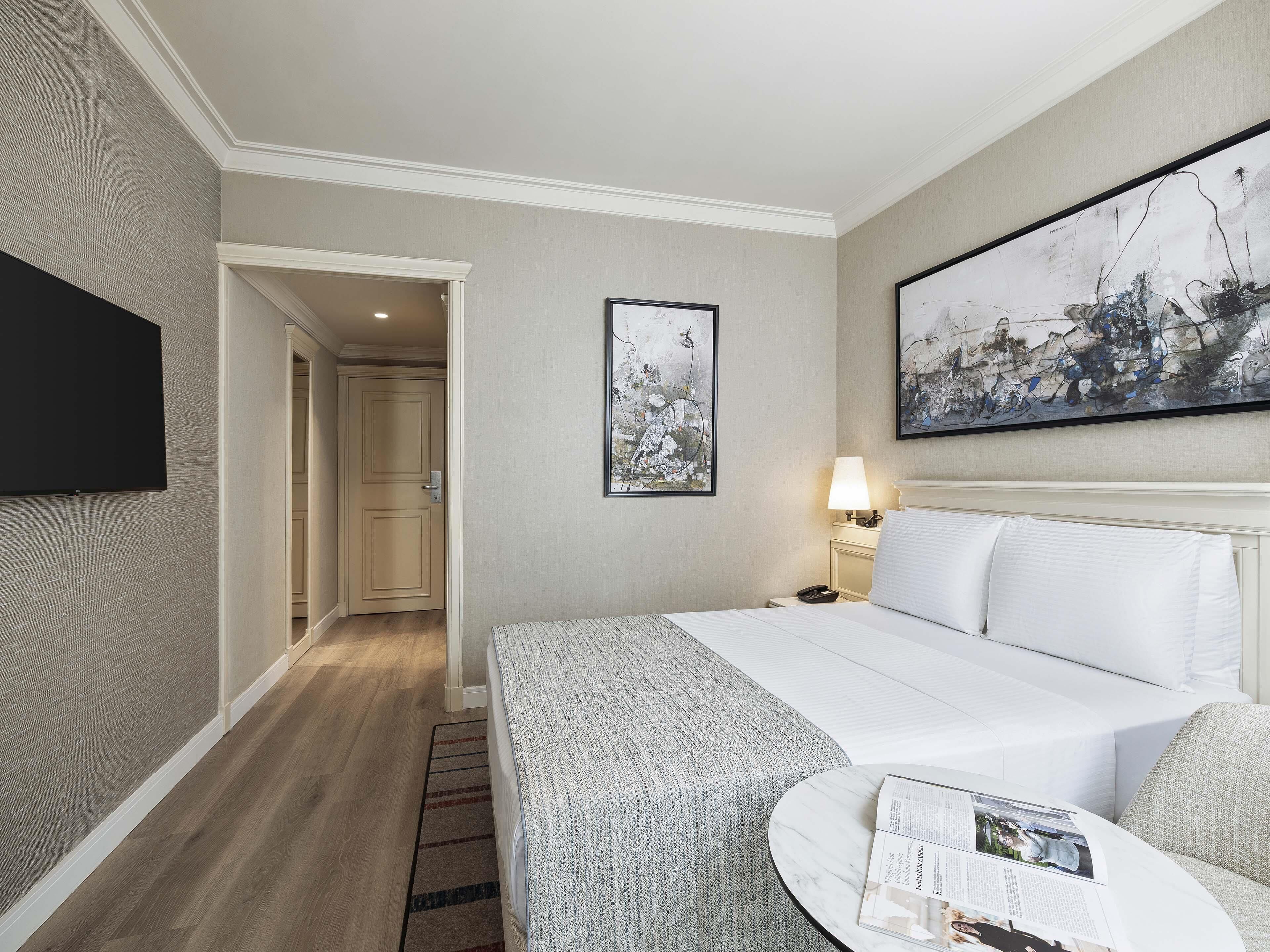 A hotel room with a bed, a bedside table with a lamp, a large abstract painting, and a television mounted on the wall.