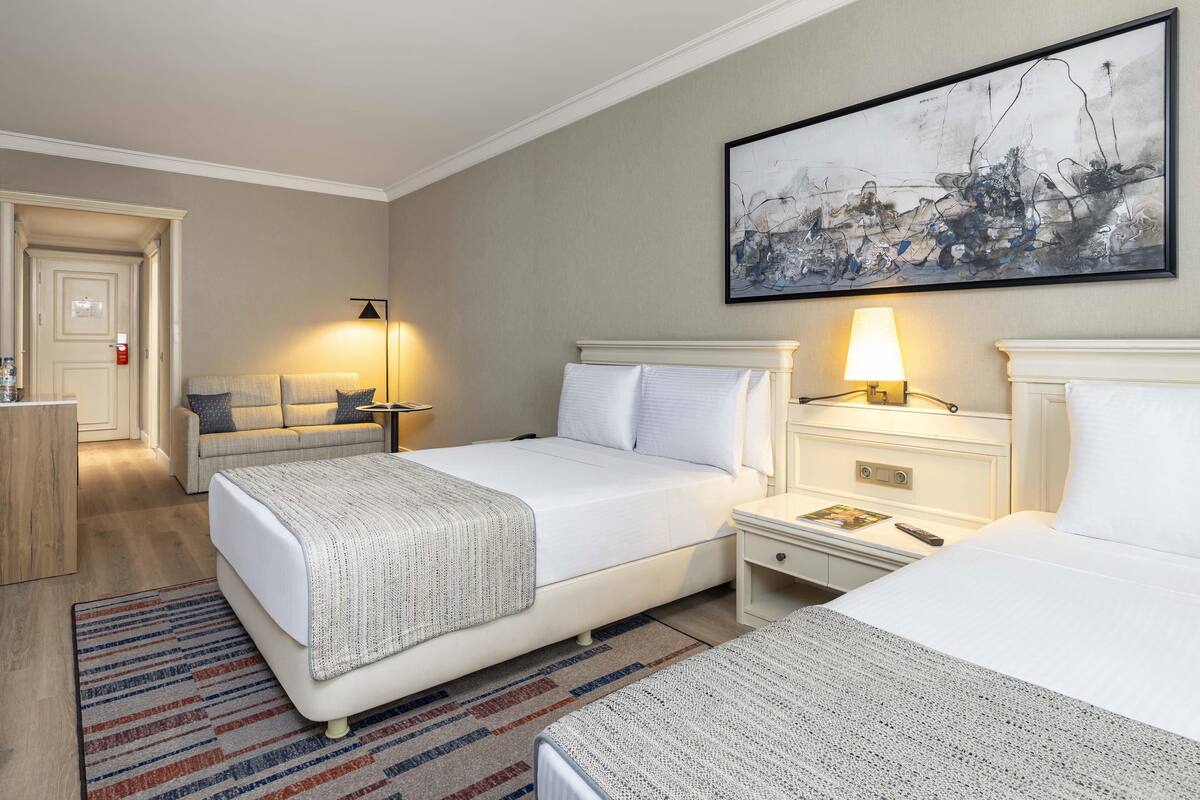 A hotel room with two beds, a large abstract painting, a bedside table with a lamp, and a small round table with a lamp.