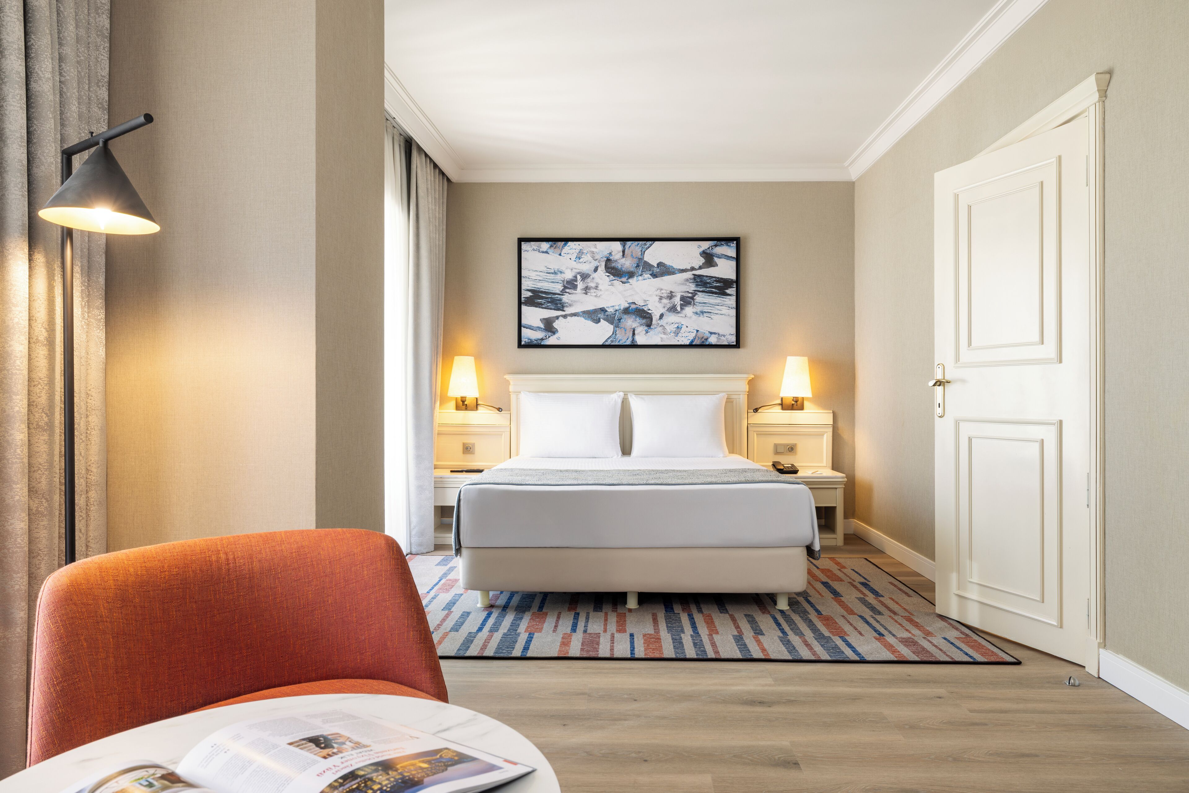 A hotel room with a bed, two bedside lamps, a framed artwork above the bed, a patterned rug, and a chair with a magazine.