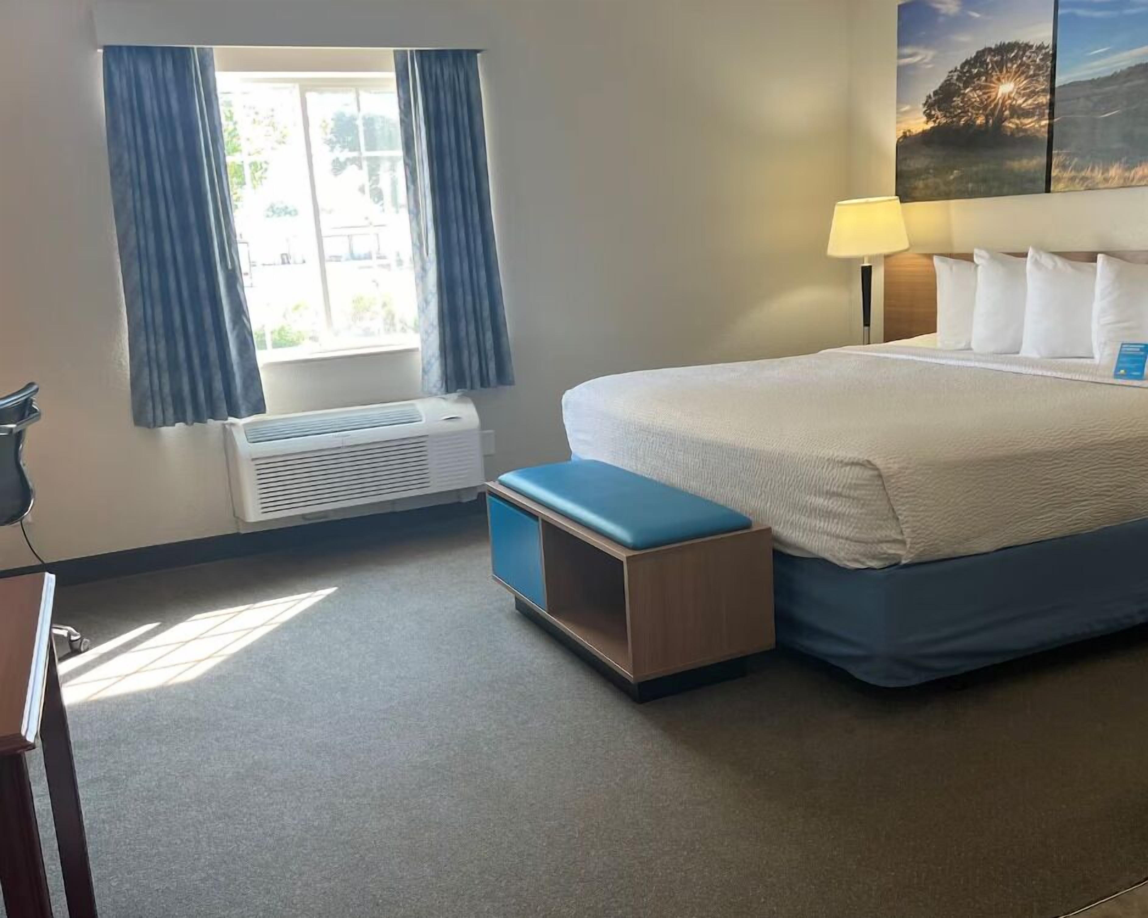Una habitación de hotel con cama, mesita de noche, lámpara, ventana con cortinas y aire acondicionado.