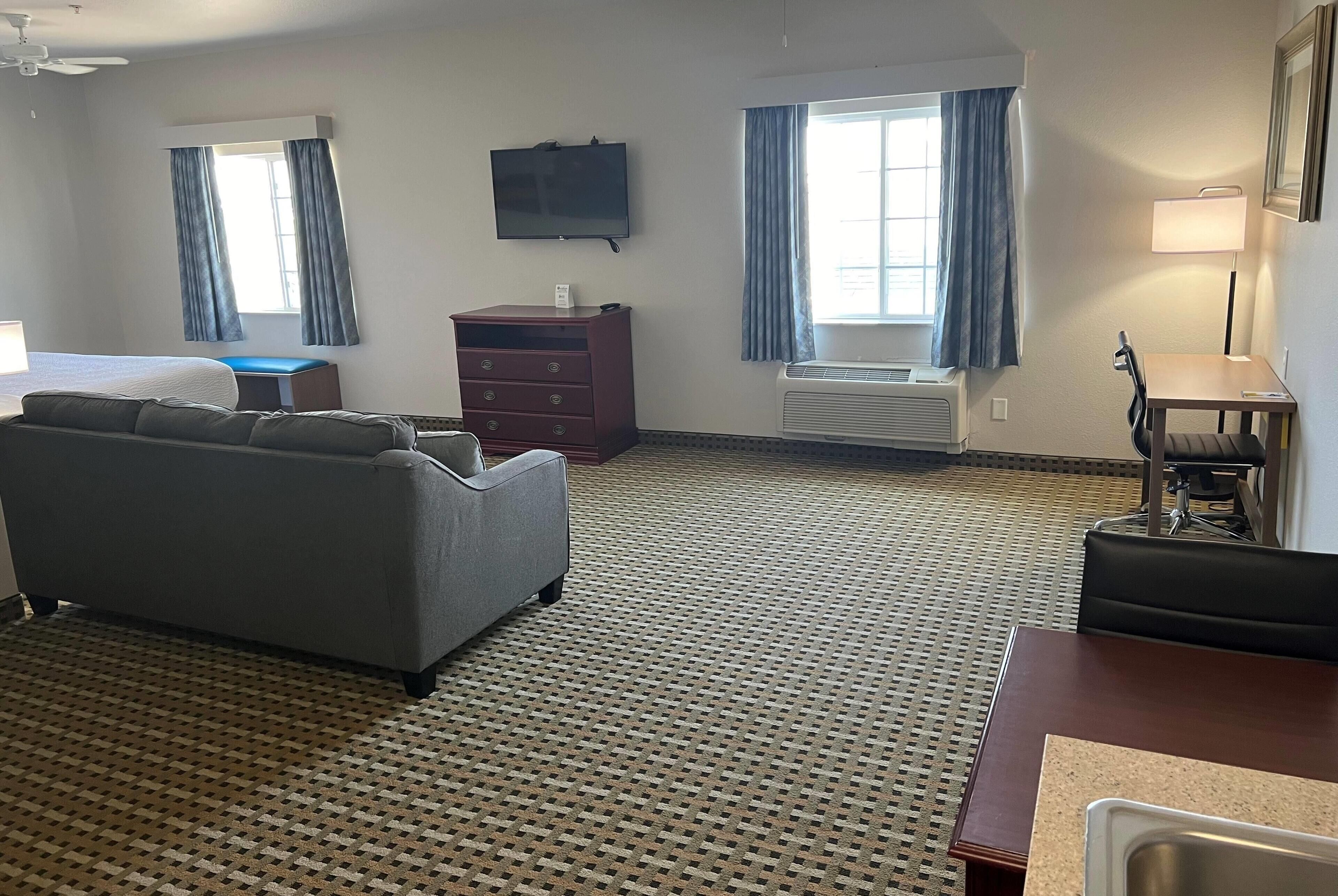 Une chambre d’hôtel avec un canapé gris, une commode en bois, un téléviseur fixé au mur et une fenêtre avec des rideaux bleus.