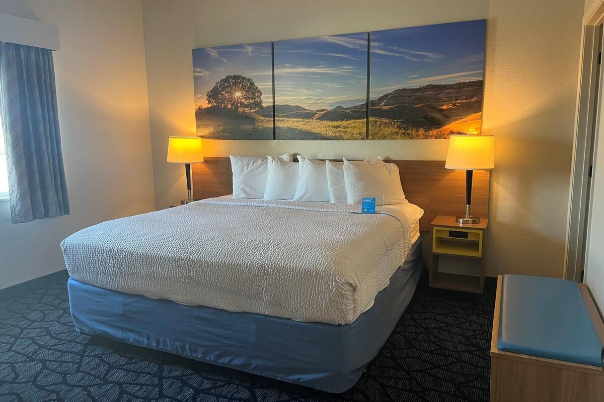 A hotel room with a large bed, two bedside lamps, a nightstand, and a view of a landscape painting.
