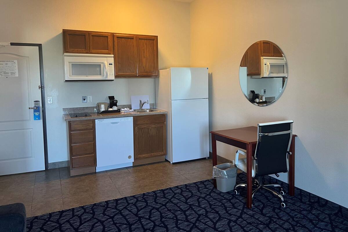 A compact kitchen with wooden cabinets, a white microwave, a white refrigerator, a dishwasher, and a small table with a chair.