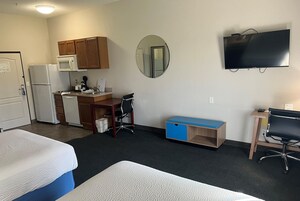 Habitación de hotel con dos camas, kitchenette, escritorio, silla, televisor y espejo.