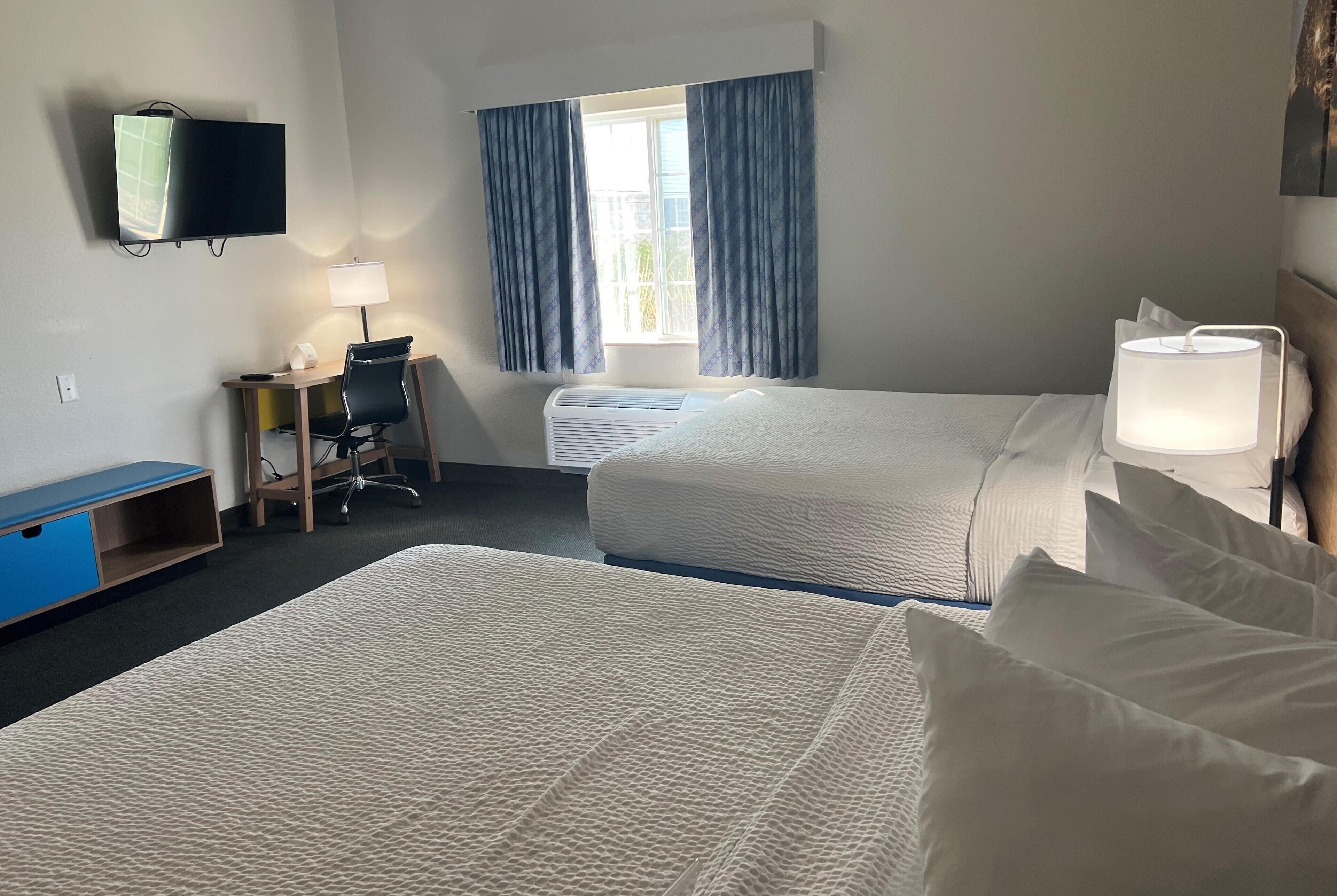 A hotel room with two beds, a desk with a chair, a TV mounted on the wall, and a window with curtains.