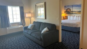 Une chambre d’hôtel avec un canapé gris, un tableau encadré au mur, un lit avec une literie blanche et une moquette à motifs.