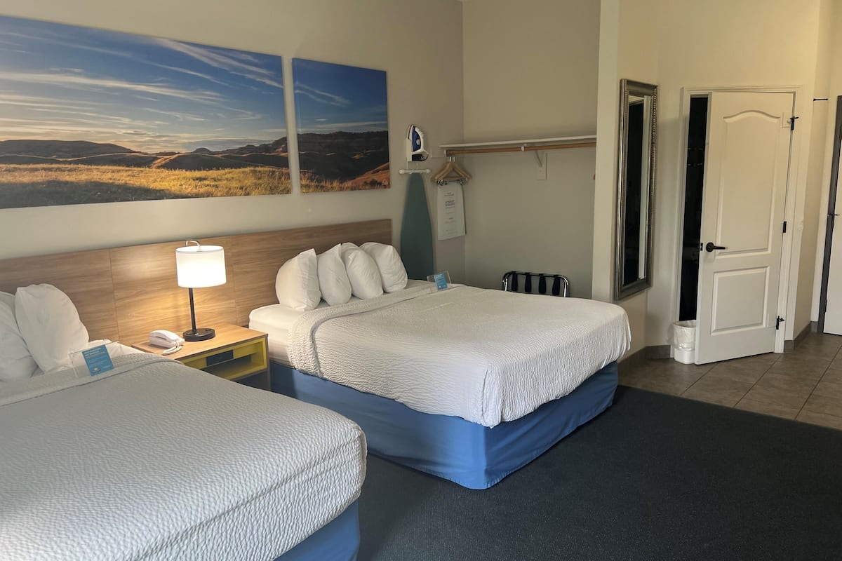 A hotel room with two beds, a nightstand with a lamp, a painting on the wall, and a door to the right.