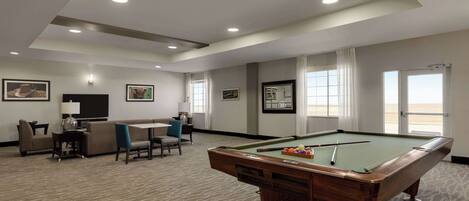 A pool table in a well-lit room with a sofa and chairs, framed pictures on the walls, and large windows.