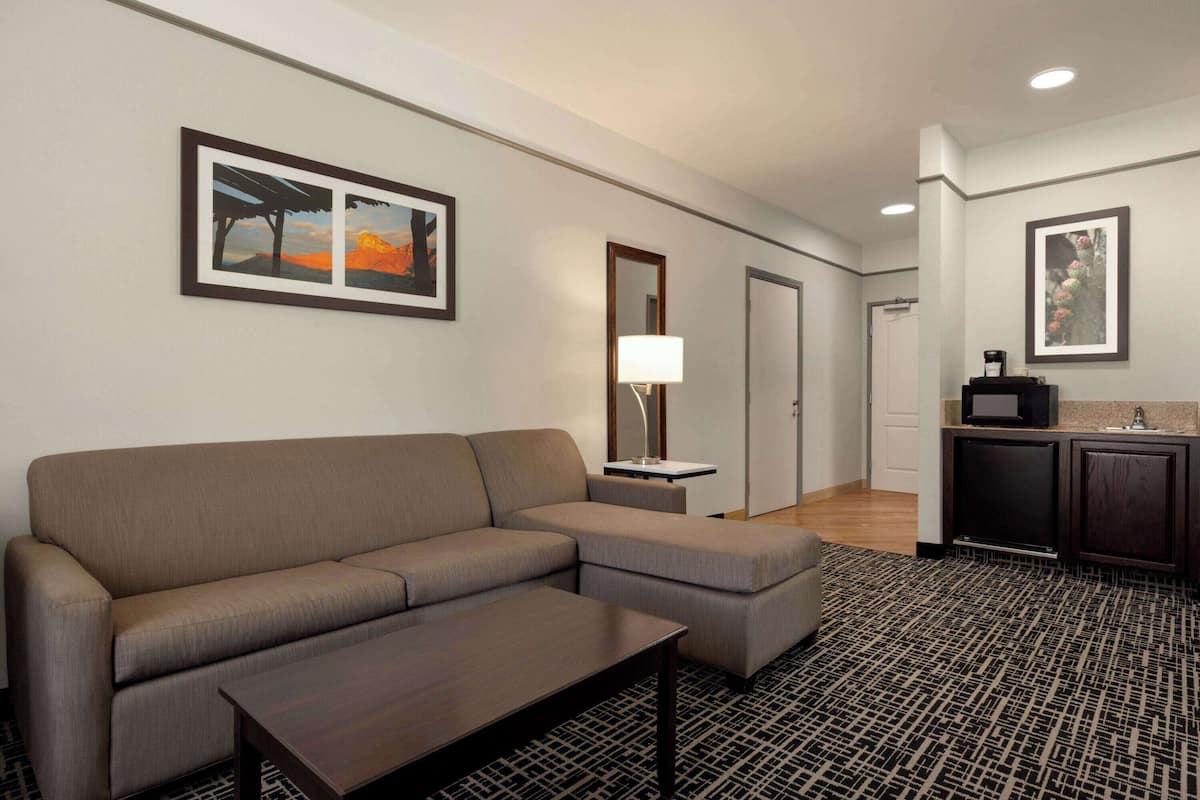 A modern hotel room with a sectional sofa, a small table, a patterned rug, a TV, and a kitchenette area.