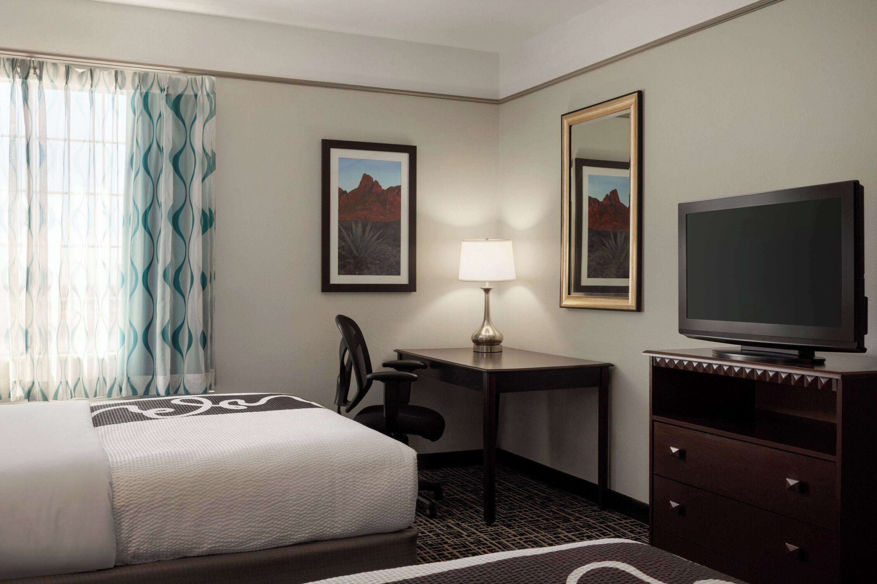 A hotel room with a bed, desk, chair, lamp, TV, and two framed pictures on the wall.