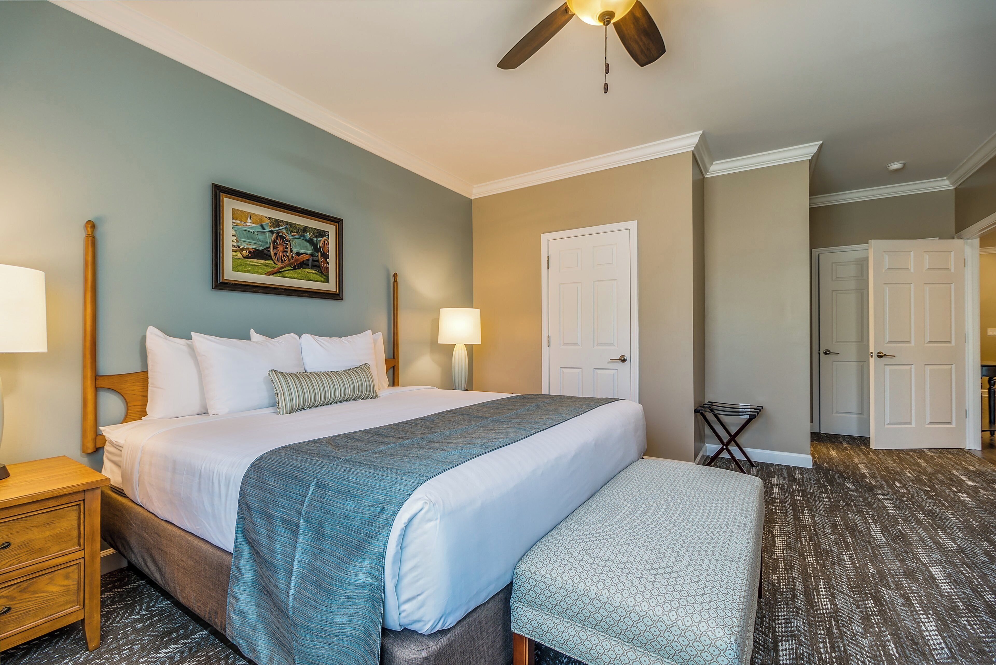 A hotel room with a bed, bedside lamps, a ceiling fan, a door, and a small table with a lamp.