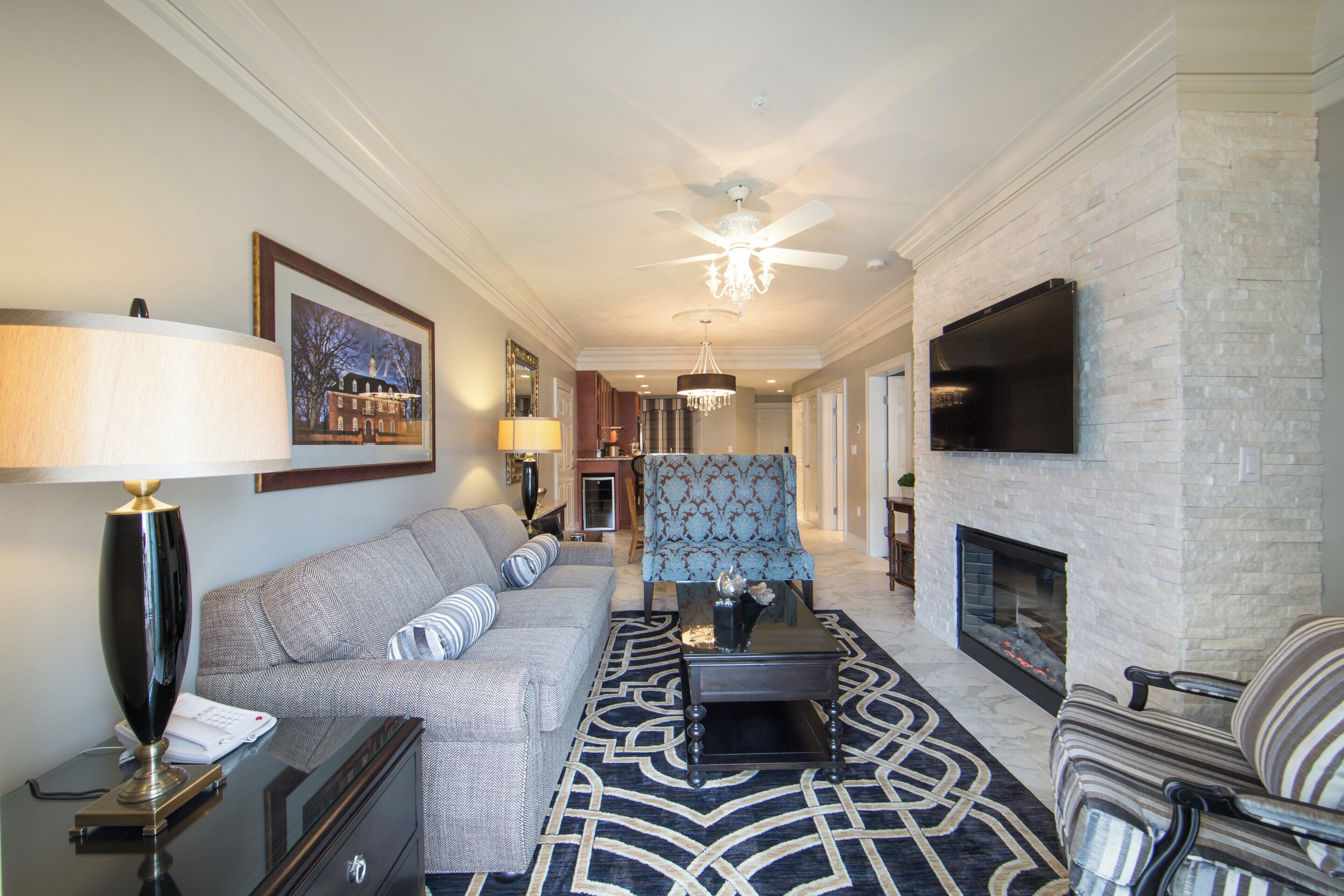 A spacious living room with a fireplace, a large flat-screen TV mounted on the wall, a ceiling fan, and a patterned rug.