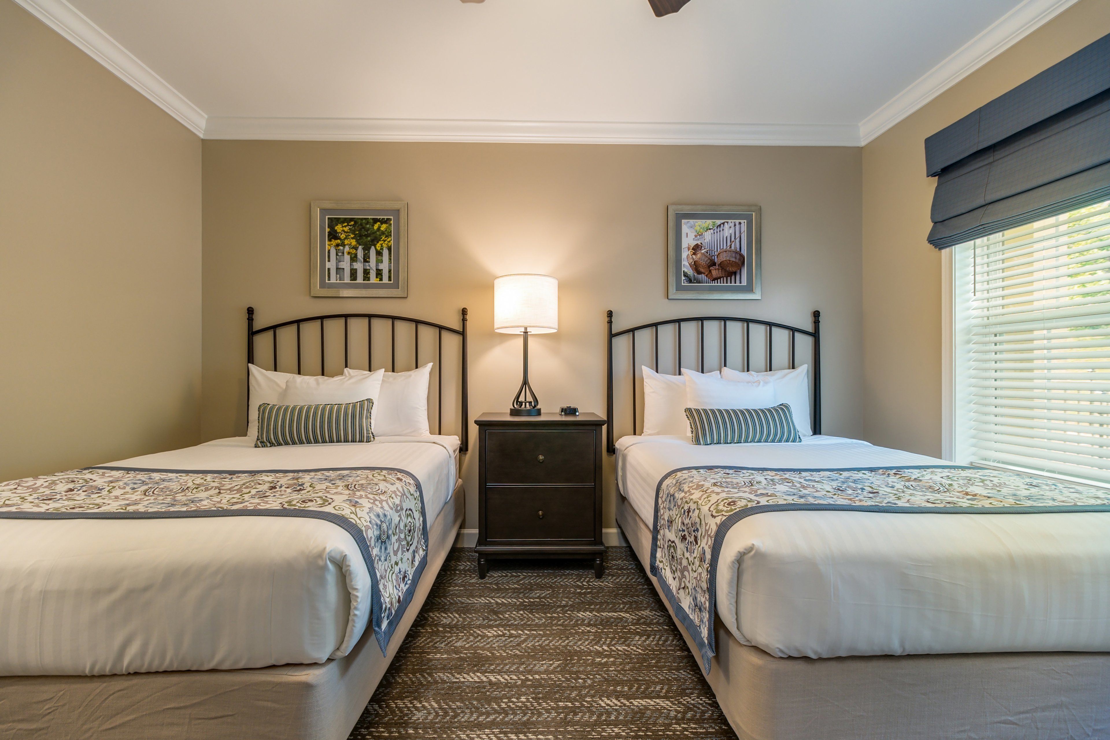 A hotel room with two beds, a nightstand with a lamp, and two framed pictures on the walls.