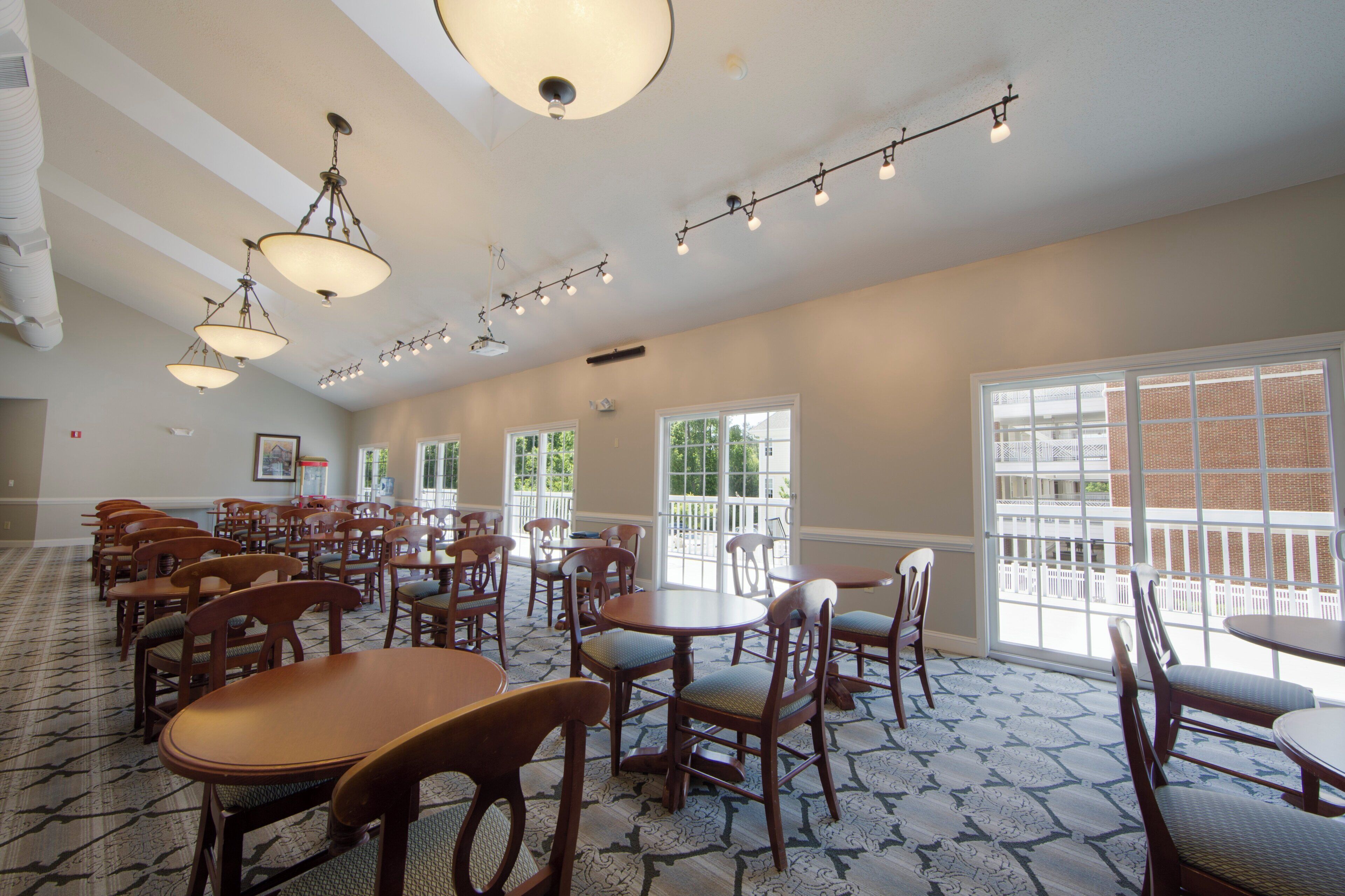 A spacious room with multiple wooden tables and chairs, large windows, and a ceiling with hanging lights.