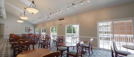 A spacious room with multiple wooden tables and chairs, large windows, and a ceiling with hanging lights.