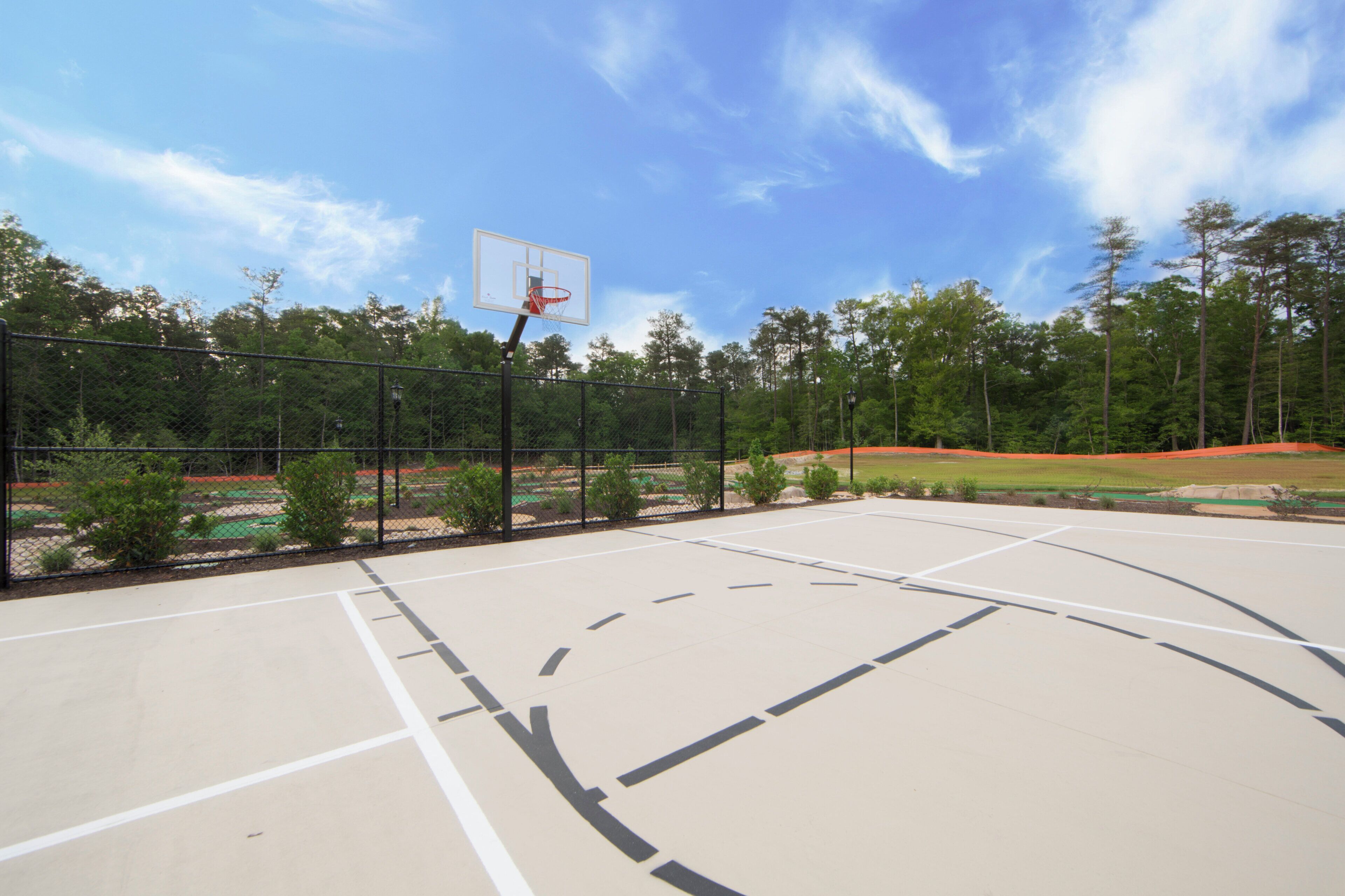 Ein Basketballplatz im Freien mit einem Maschendrahtzaun, Bäumen und blauem Himmel.