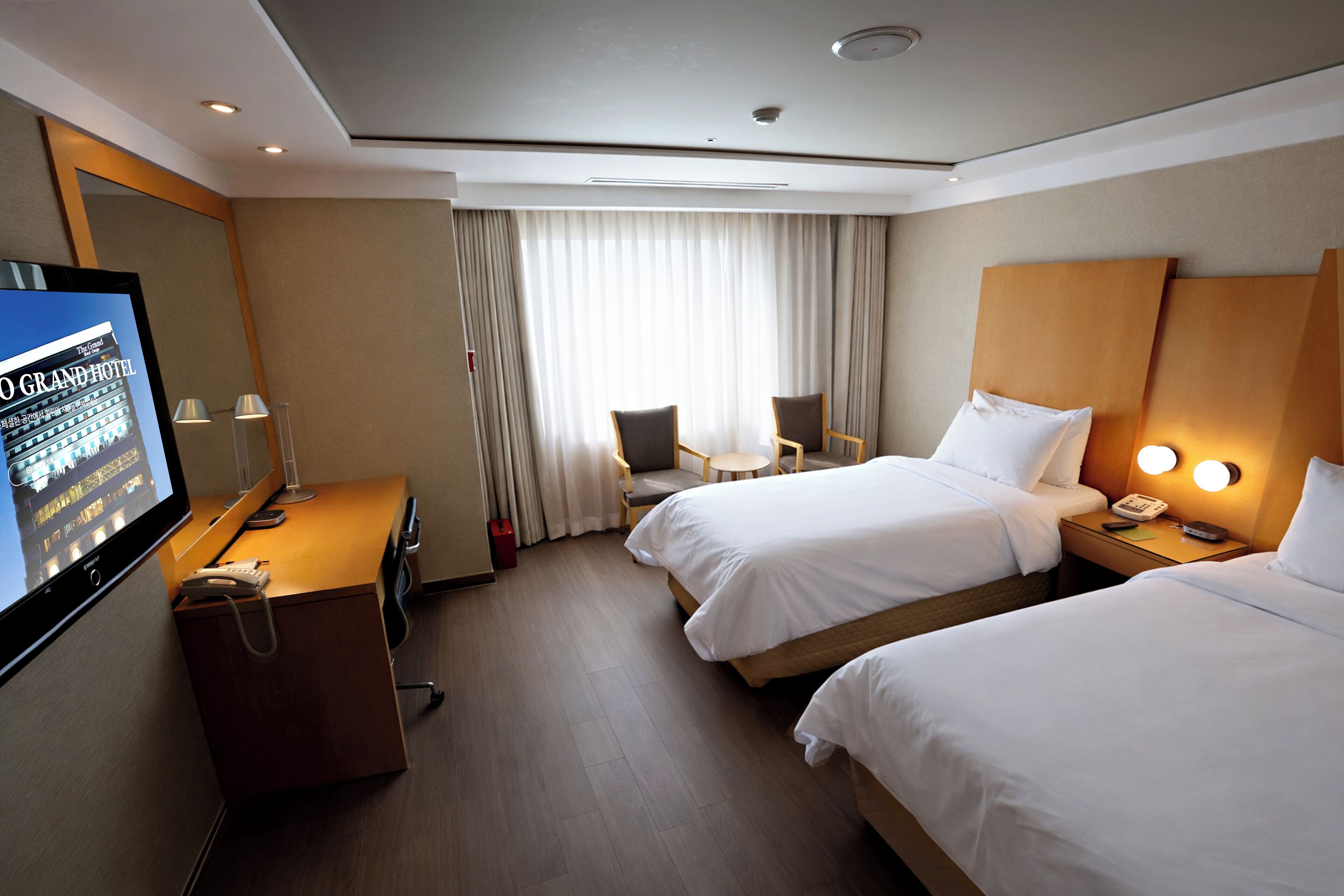 A hotel room with two beds, a desk, a TV, and a window with curtains.