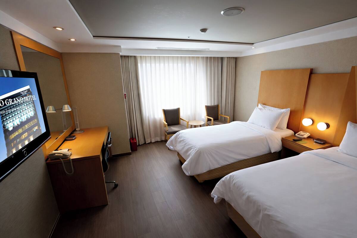 A hotel room with two beds, a desk, a TV, and a window with curtains.