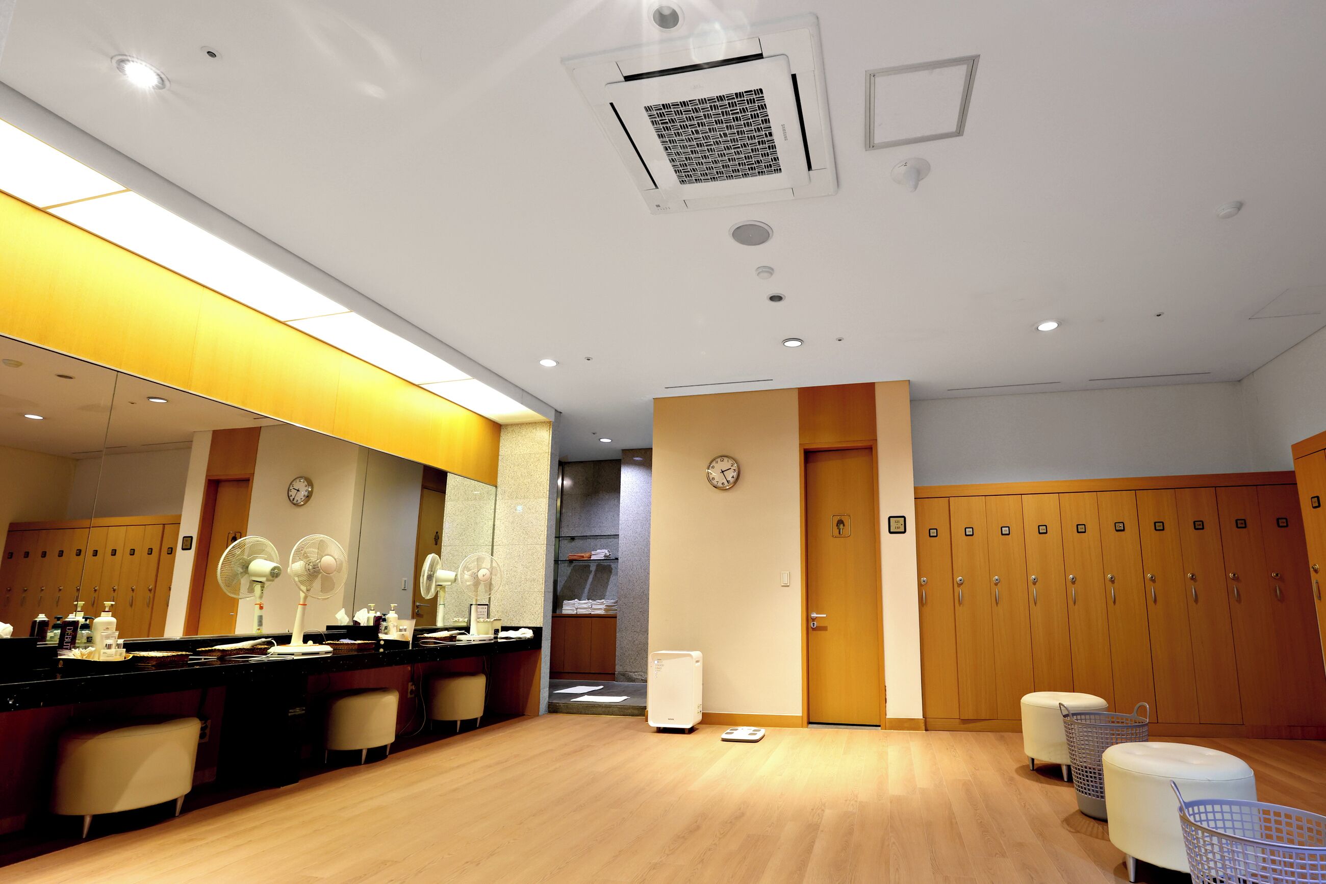 A hotel room with a wooden floor, a mirror, a desk with a chair, a scale, and a wardrobe with multiple compartments.