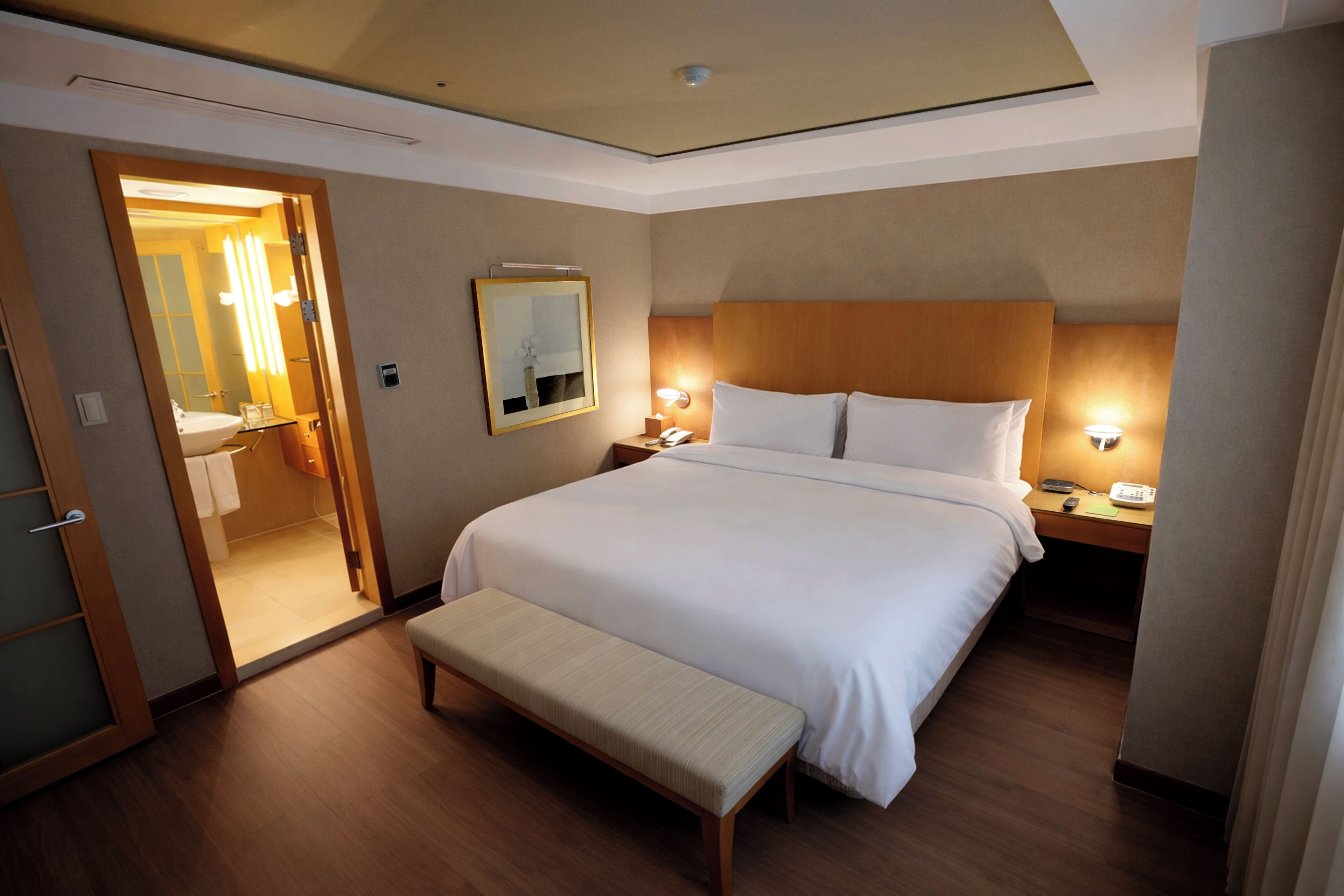 A hotel room with a large bed, bedside tables, a bench, a mirror, and a bathroom visible through an open door.