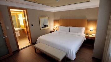 Habitación de hotel con una cama grande, mesitas de noche, un banco, un espejo y un baño visible a través de una puerta abierta.
