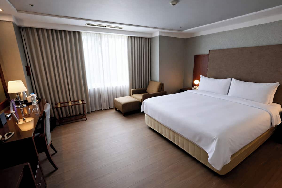 Quarto de hotel com uma cama grande, uma escrivaninha com luminária, uma cadeira e uma janela com cortinas.