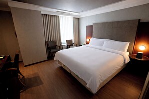 A hotel room with a large bed, a desk with a chair, a small table, and a lamp.