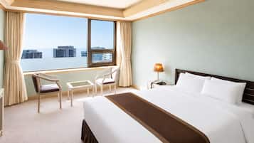 Habitación de hotel con una cama grande, un escritorio con televisor, una silla, una mesita, una lámpara y una ventana con vistas a edificios.