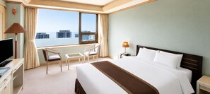 A hotel room with a large bed, a desk with a TV, a chair, a small table, a lamp, and a window with a view of buildings. - Libero Hotel (Busan)