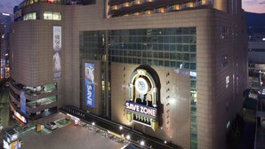 Een meerhoog hotel met een opvallend 'SAVEZONE' bord, een klok en een 'LOTTERY' winkel op de begane grond.