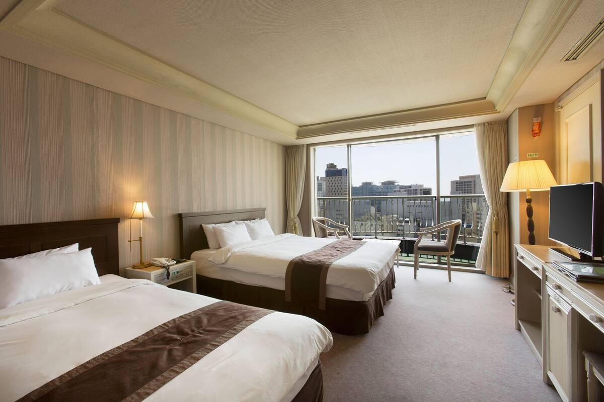 A hotel room with two beds, a desk with a TV, a chair, and a large window offering a city view.