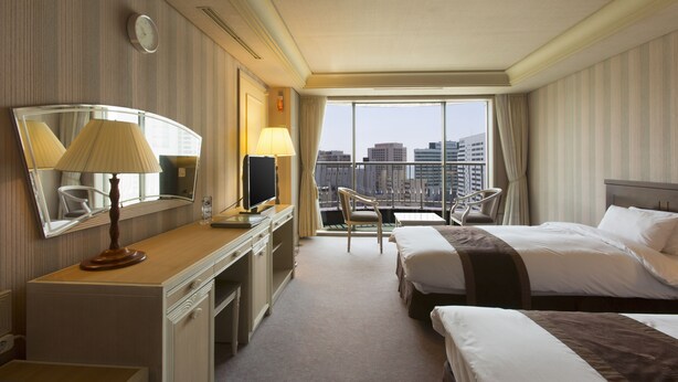 Een hotelkamer met een groot bed, een kleiner bed, een bureau met een lamp, een stoel en een groot raam met uitzicht op de stad.
