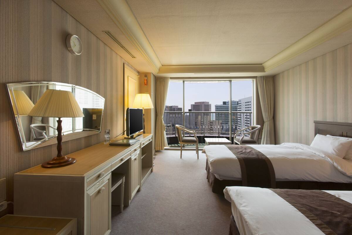 A hotel room with a large bed, a smaller bed, a desk with a lamp, a chair, and a large window offering a city view.