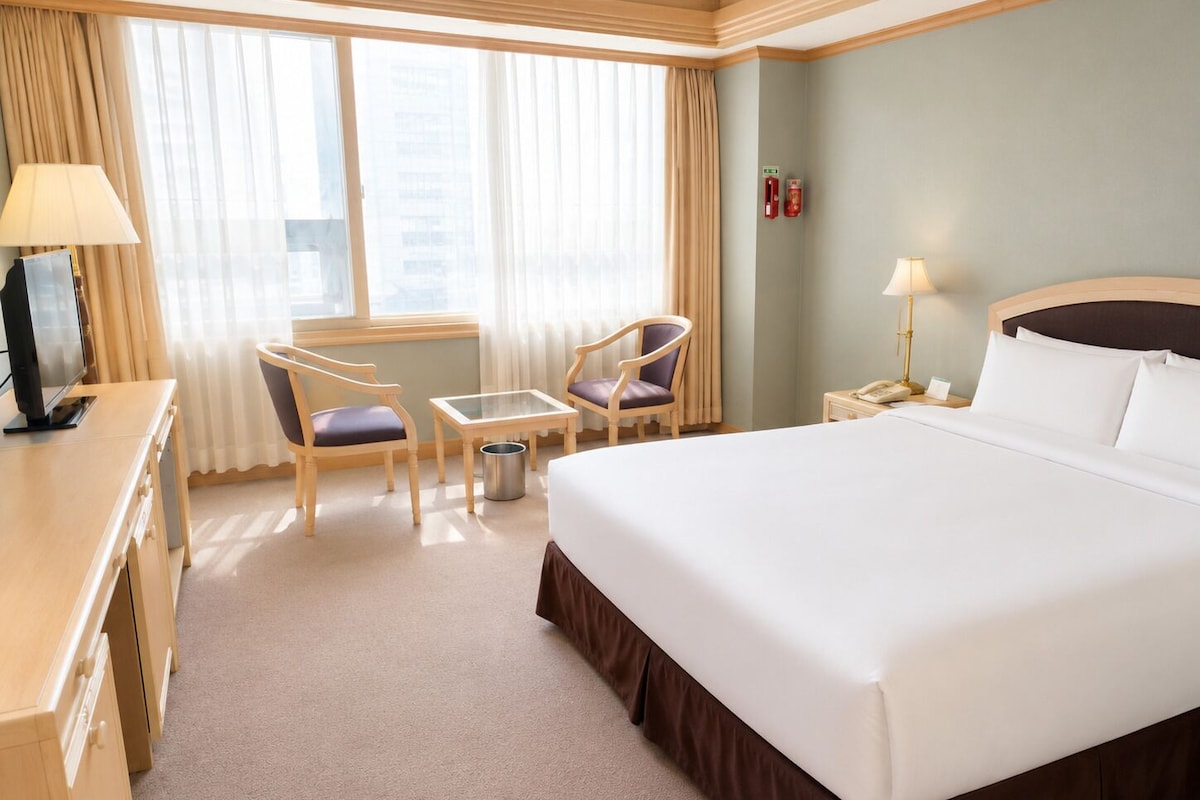 Une chambre d’hôtel avec un grand lit, un bureau, deux chaises, une petite table, une lampe, un téléphone et une fenêtre avec des rideaux.