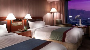 A hotel room with two beds, nightstands, lamps, a chair, and a view of a cityscape at dusk. - Hotel Paragon (Busan)