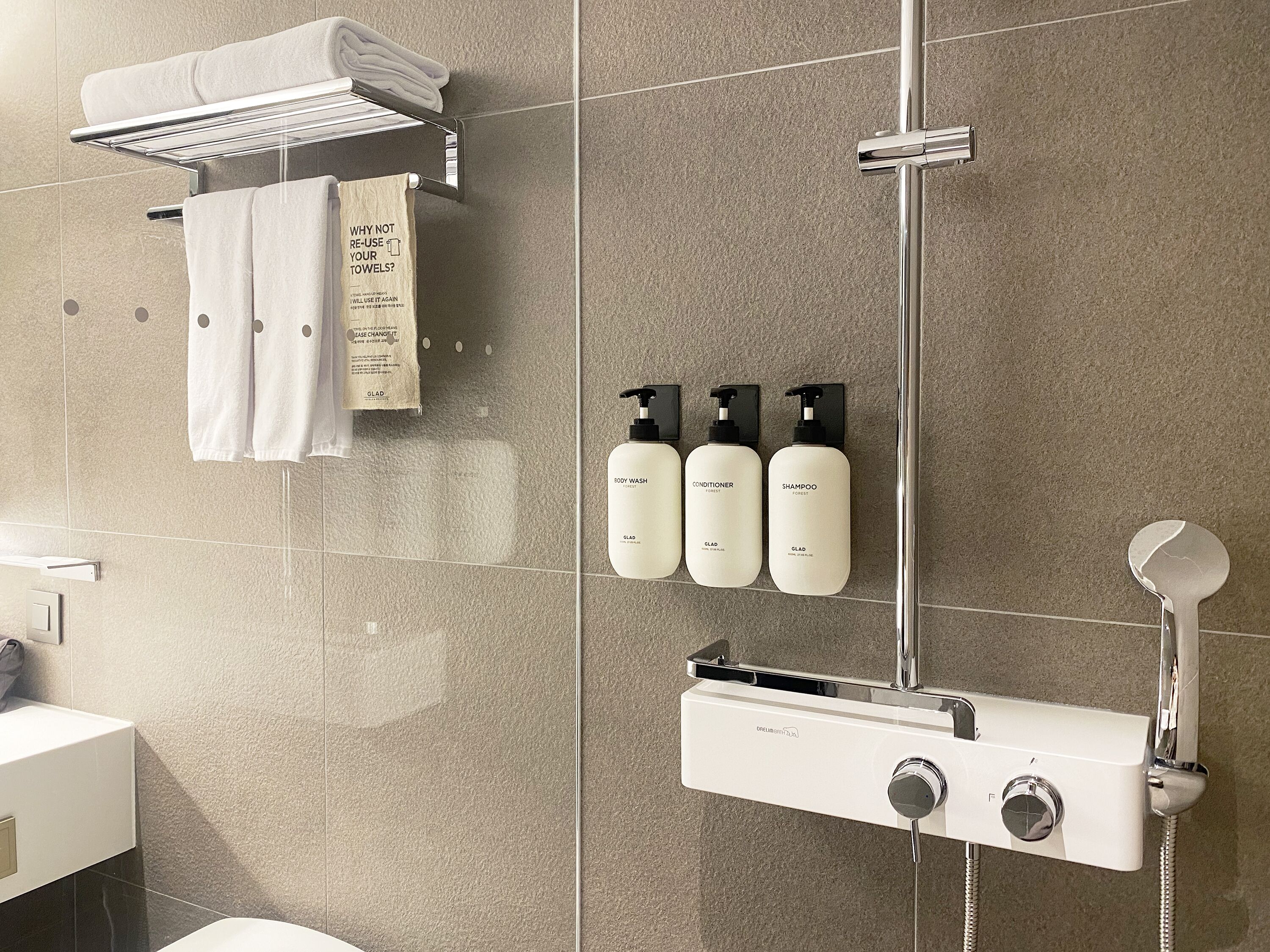 A modern bathroom with a shower, a towel rack with folded towels, and three toiletries (body wash, conditioner, shampoo) mounted on the wall.