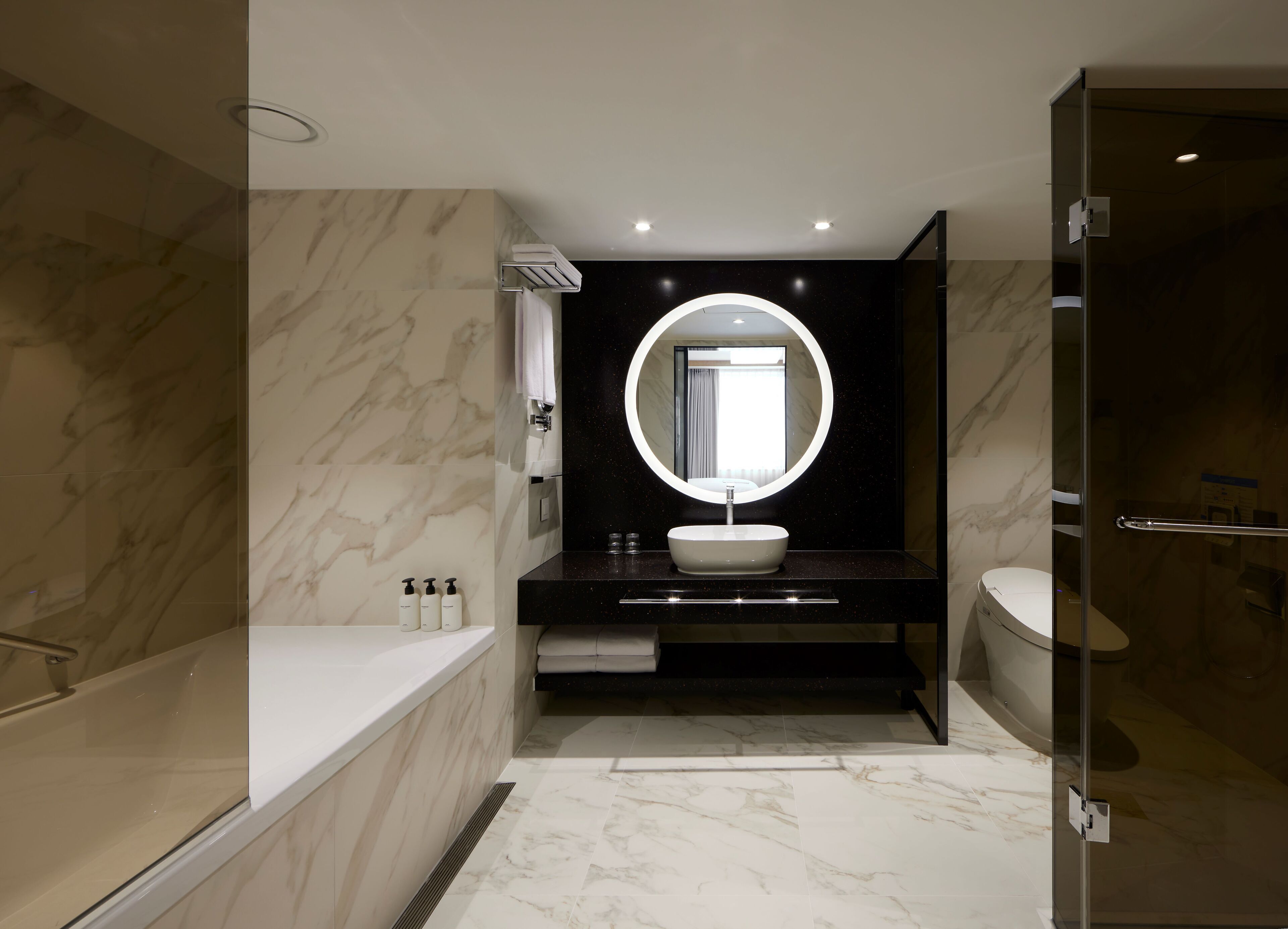 Un baño moderno con suelo de mármol, una bañera independiente, un espejo redondo y un box de ducha de vidrio.