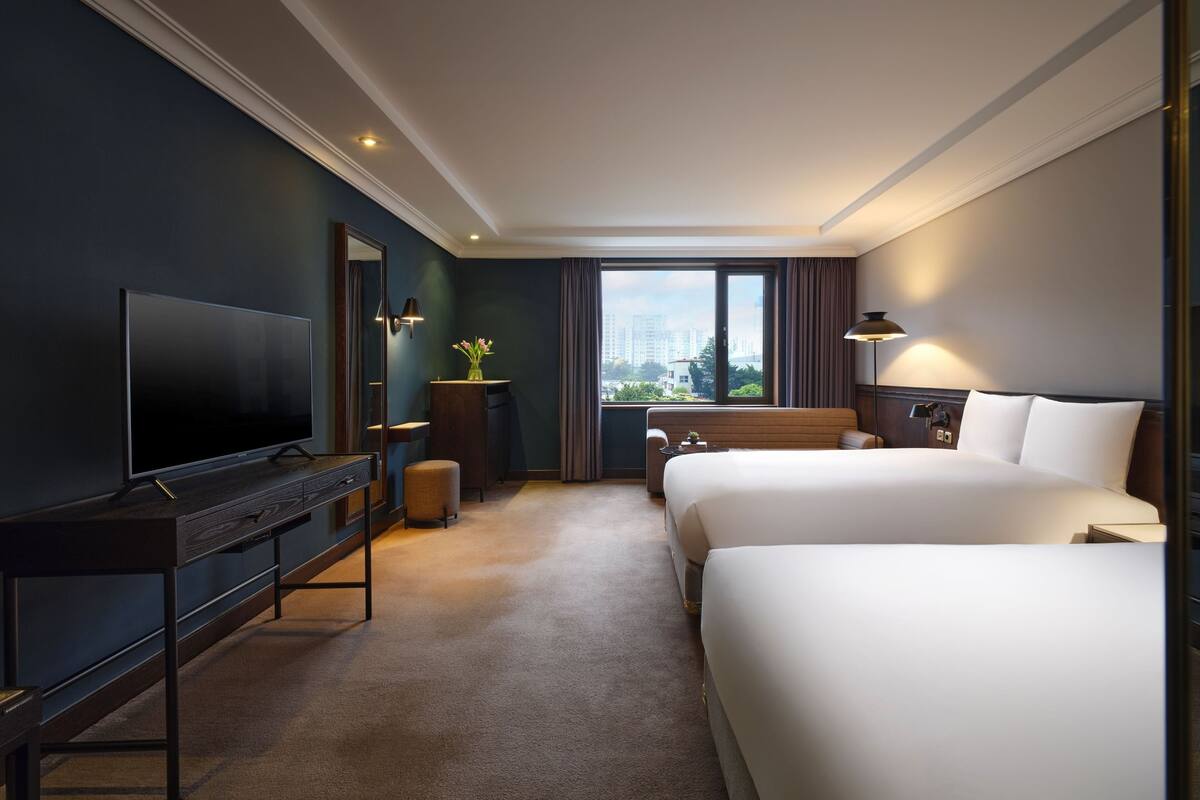 Een hotelkamer met een groot flatscreen televisietoestel, een bureau met lades, een bank, twee bedden, een raam met gordijnen en uitzicht op gebouwen.