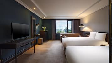 Habitación de hotel con una televisión de pantalla plana grande, un escritorio con cajones, un sofá, dos camas, una ventana con cortinas y vistas a edificios.