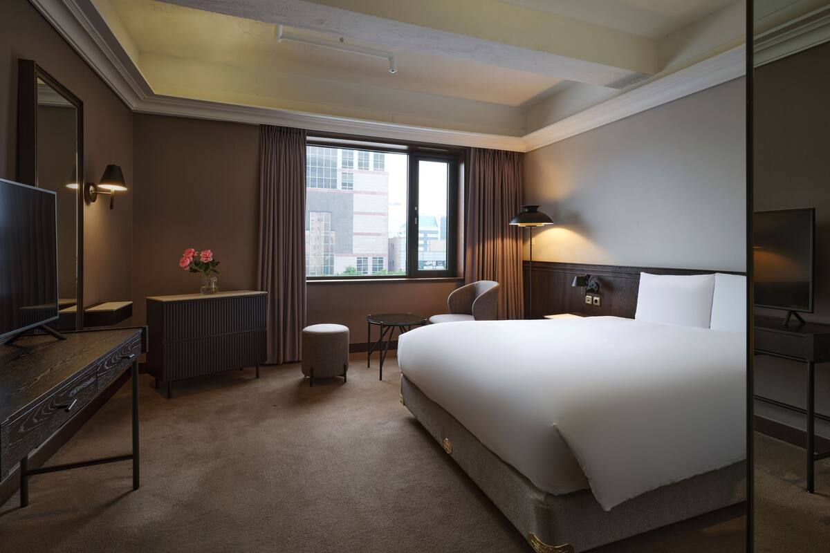 Een hotelkamer met een groot bed, een bureau met spiegel, een stoel, een radiator, een raam met gordijnen en een televisie.