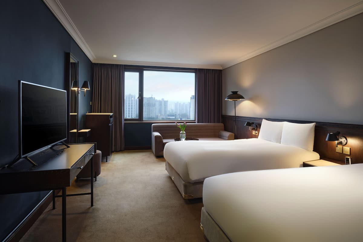 Een hotelkamer met een groot bed, een kleiner bed, een bureau met een televisie, een stoel, een lamp en uitzicht op de stad.